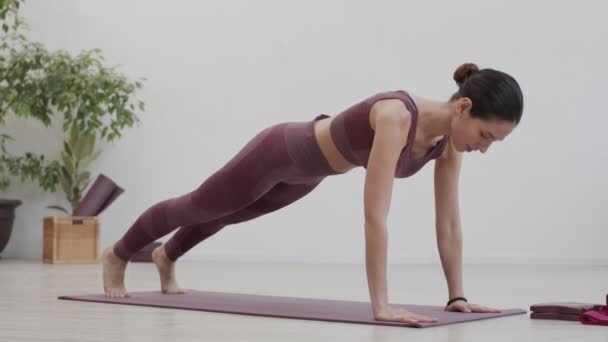 Amplio tiro de mujer atlética joven caucásica usando ropa deportiva haciendo ejercicios de yoga en la estera de yoga en el interior - Imágenes, Vídeo