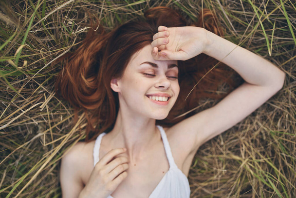 Ευτυχισμένη γυναίκα ξαπλώνει στο σανό έξω με ένα λευκό φόρεμα και χειρονομίες με τα χέρια της - Φωτογραφία, εικόνα