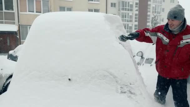 Śnieg i burze. Mężczyzna czyści pokryty śniegiem samochód. Duże opady śniegu. Kierowca w plecaku ze szczotką usuwa śnieg z samochodu stojącego na parkingu - Materiał filmowy, wideo