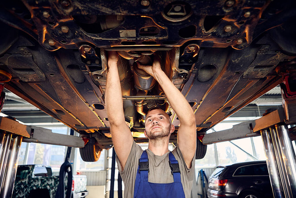 Ansehnlicher Automechaniker, der das Fahrwerk des Autos an der Tankstelle überprüft. Fröhliche männliche Arbeiter reparieren Problem mit dem Auto. Konzept der Problemlösung - Foto, Bild
