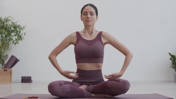 Bloqueio de jovens caucasianas yogi feminino vestindo sportswear sentado na posição de lótus no tapete de ioga dentro de casa, meditando e olhando para a câmera - Filmagem, Vídeo