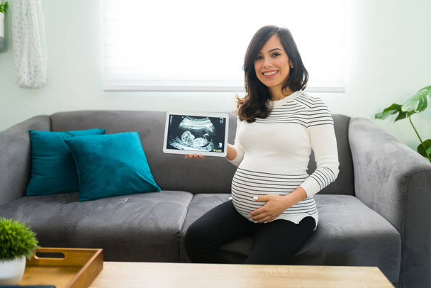 Mujer embarazada feliz sonriendo y sosteniendo una tableta con el ultrasonido digital de su nuevo hijo en la pantalla mientras está sentada en su sofá de la sala de estar  - Foto, Imagen