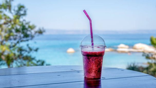 Cocktail läpinäkyvässä muovikupissa oljilla ja näkymä Egeanmeren rannikolle taustalla, Kreikka - Valokuva, kuva