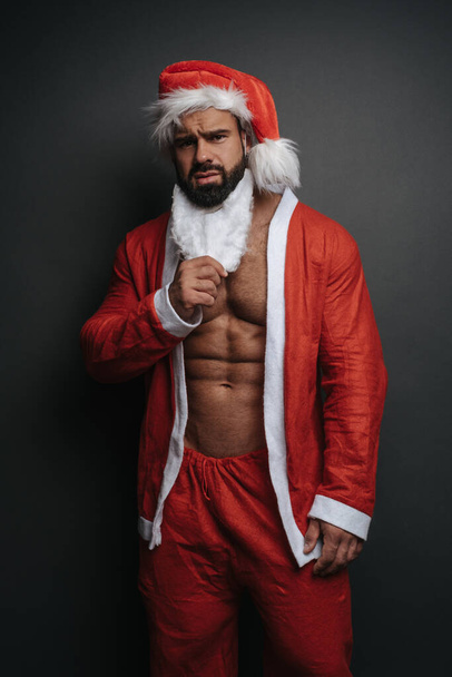 スタジオの灰色の背景でサンタハットの筋肉質の男を髭を下に移動させます。クリスマスのハンサムな男が見える。セクシーフックで赤サンタクロースの衣装. - 写真・画像