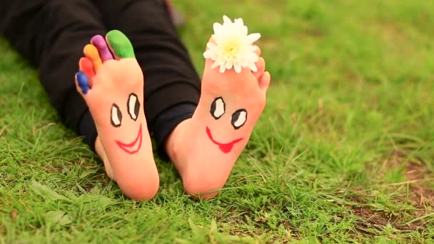 Маленькая девочка сидит на траве с раскрашенными ногами и смешными комическими лицами  - Кадры, видео
