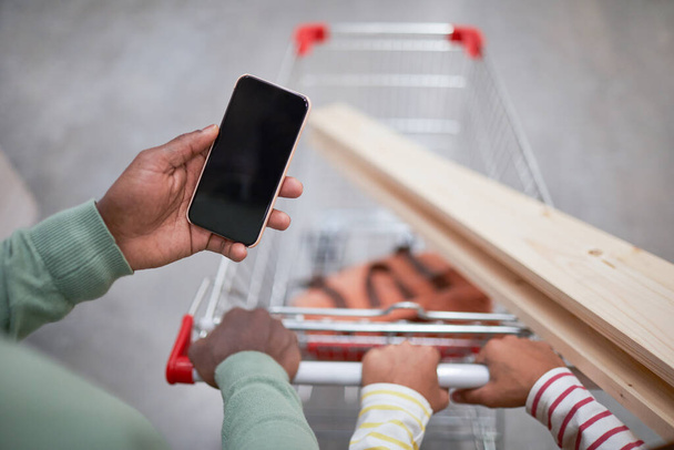 Primer plano de un hombre afroamericano irreconocible sosteniendo un teléfono inteligente con pantalla en blanco mientras empuja el carrito de compras en el supermercado, espacio para copiar - Foto, imagen