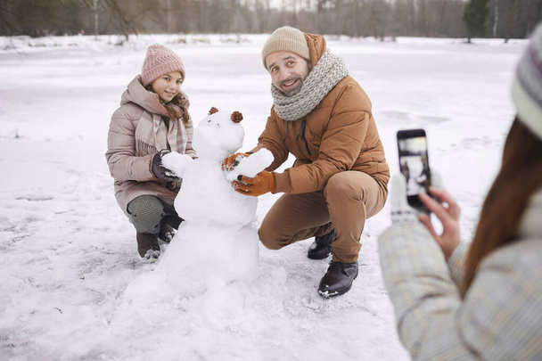 Portret młodej kobiety robiącej zdjęcia ojcu i córce budującej bałwana razem podczas zimy na świeżym powietrzu, przestrzeń kopiująca - Zdjęcie, obraz