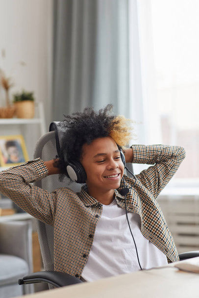 Evde dizüstü bilgisayar, oyuncu ya da blogger konsepti kullanırken koltukta dinlenen ve kulaklık takan gülümseyen Afro-Amerikalı çocuğun dikey portresi - Fotoğraf, Görsel