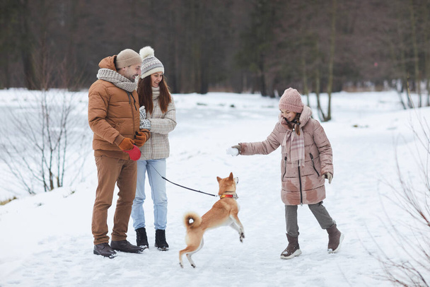Full length portrait of happy family playing with dog outdoors while enjoying walk in winter forest by lake, focus on cheerful girl wearing pink jacket holding snowball, copy space - Photo, Image