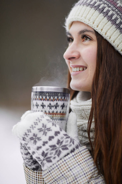 Vue latérale verticale portrait d'une femme adulte souriante tenant une tasse de chocolat chaud et regardant ailleurs tout en profitant de la promenade en plein air en hiver - Photo, image