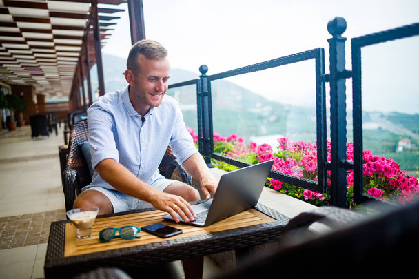 Un uomo d'affari di successo è seduto a un tavolo con un computer portatile, sta lavorando in vacanza. Vacanza e lavoro a distanza. Il ragazzo è seduto in un ristorante su una terrazza con vista panoramica - Foto, immagini