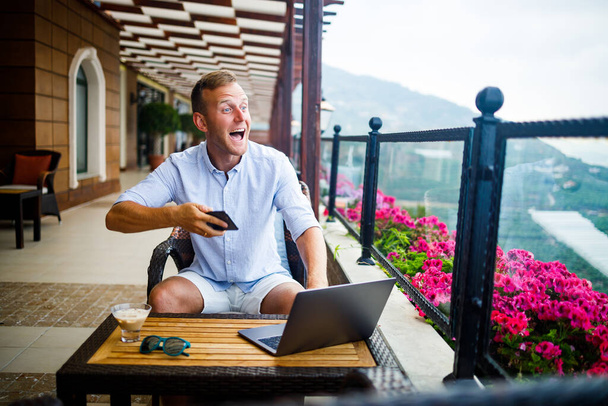 Joven hombre de negocios guapo en una camisa blanca y pantalones cortos se sienta con un ordenador portátil en un café a la mesa. Trabajo mientras descansa - Foto, imagen