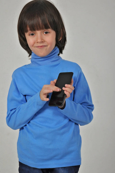 Mobile app for children. Happy excited cheerful little boy with dark hair pointing at cell phone and smiling at camera, showing telephone. indoor studio shot isolated on grey background - Photo, Image