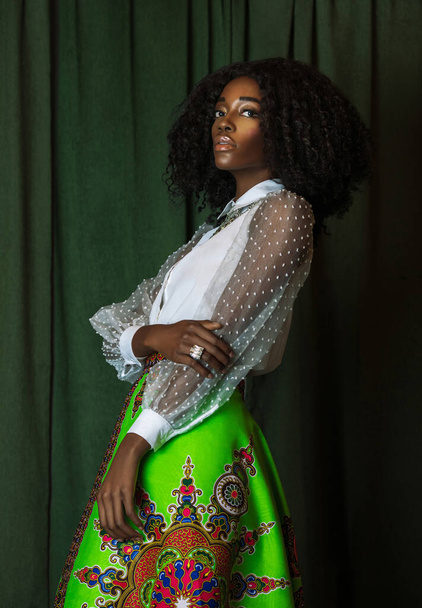 Portrait of an sexy black woman with curly long black hair, beautiful makeup and soft lips posing by herself in a studio with green curtain background wearing a white blouse and colorful skirt. - Photo, Image