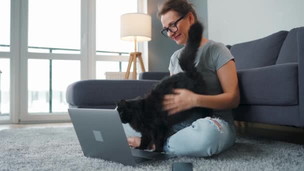 Femme vêtue occasionnellement avec des lunettes s'assoit sur un tapis avec un ordinateur portable et caresse un chat noir moelleux tout en travaillant dans une chambre confortable. - Séquence, vidéo