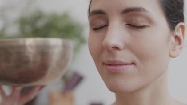 Close up of young Caucasian woman meditating with closed eyes while listening to sounds of yoga singing bowl unrecognizable woman holding and moving on background - Footage, Video
