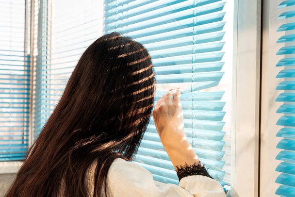 Achteraanzicht - een vrouw met lang haar gluurt uit het raam door de gordijnen van blauwe jaloezieën - Foto, afbeelding