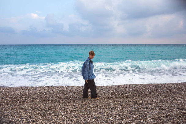 Una ragazza con i capelli rossi corti cammina lungo la spiaggia, un hipster, una donna cammina lungo il mare, una ragazza con una camicia lunga e pantaloni larghi - Foto, immagini