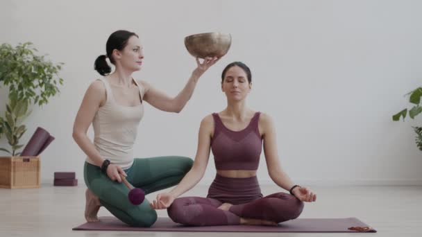 Lezárása nő sztrájk jóga éneklő tál és elkezd mozgatni a kezét, míg a fiatal kaukázusi sportnő ül jóga mat csukott szemmel és meditál - Felvétel, videó