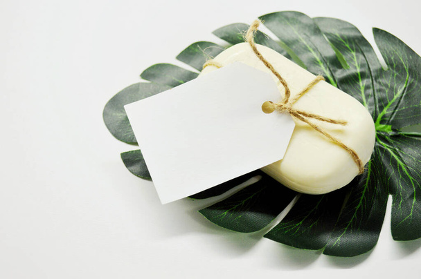 Λευκό σαπούνι δεμένο με σχοινί με ετικέτα βρίσκεται σε λευκό φόντο σε ένα πράσινο φύλλο φοίνικα - Φωτογραφία, εικόνα