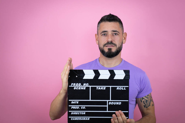 Young handsome man wearing casual t-shirt over pink background holding clapperboard very happy having fun - Photo, Image