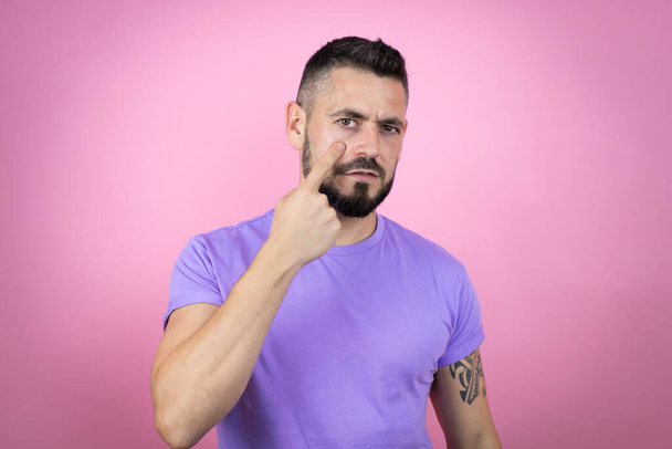 Fiatal jóképű férfi alkalmi pólót visel rózsaszín háttér mutatva a szem figyeli gesztus, gyanús kifejezés - Fotó, kép