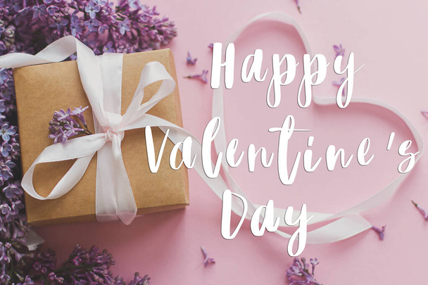 Happy Valentine's day text on lilac flowers, gift box and heart ribbon on pink paper, flat lay. Love concept. Valentines day stylish greeting card - Photo, Image