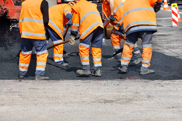 Ein Team von Straßenarbeitern in orangefarbenen reflektierenden Uniformen bedeckt einen Straßenabschnitt mit frischem Asphalt, um den Straßenabschnitt zu reparieren. Klarer Tag, Kopierraum. - Foto, Bild