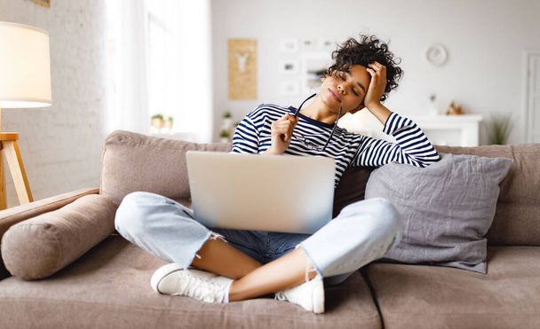 κουρασμένη εθνοτική γυναίκα με γυαλιά αγγίζοντας το κεφάλι και κλείνοντας τα μάτια αναπαύεται ενώ κάθεται σταυροπόδι κοντά στο laptop και εργάζονται σε απομακρυσμένο έργο στο άνετο σαλόνι στο σπίτι - Φωτογραφία, εικόνα