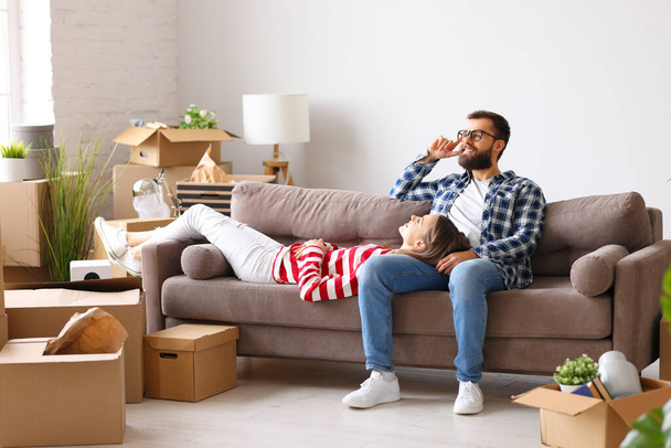 Ευτυχισμένο νεαρό ζευγάρι απολαμβάνει νέο διαμέρισμα και κάνει σχέδια ενώ στηρίζεται σε καναπέ στο δωμάτιο με συσκευασμένα κουτιά με πράγματα - Φωτογραφία, εικόνα