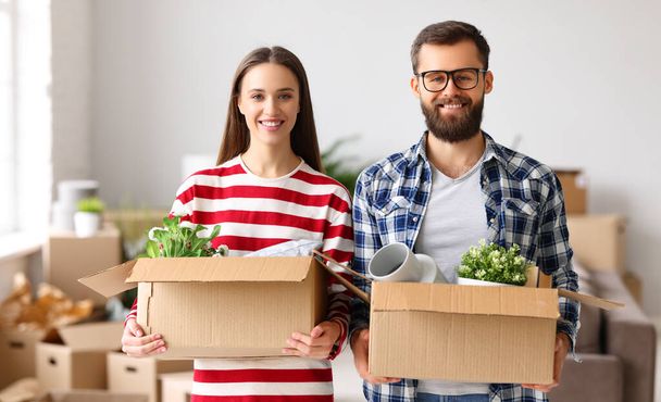 Счастливые молодой человек и женщина улыбаются и смотрят в камеру, неся коробки с вещами во время переезда в новую квартиру - Фото, изображение