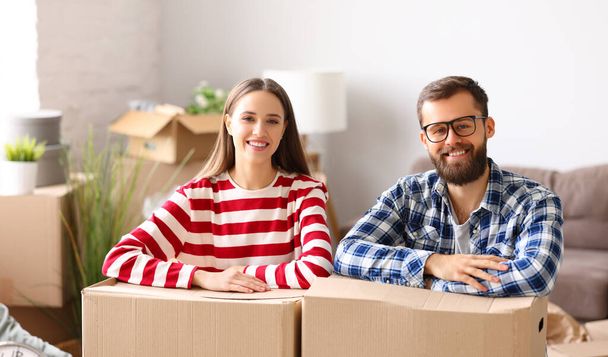 Happy young man and woman smiling and looking at camera with carton boxes with belongings during relocation into new apartment - Photo, Image