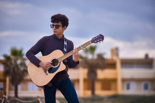 jeune adolescent caucasien jouant de la guitare acoustique sur la plage. lunettes de soleil et vêtements sombres. - Photo, image