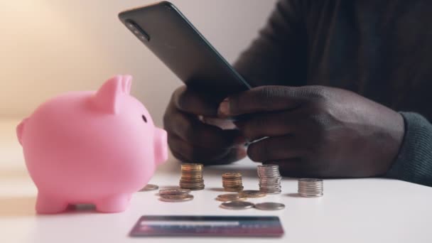 Mann benutzt Telefon, um sein Bankkonto zu überprüfen. Sparschwein-Kreditkarte und Münzen auf dem Tisch - Filmmaterial, Video