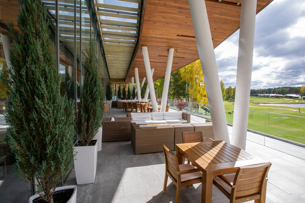 Ξύλινο τραπέζι και καρέκλες, καναπέδες και πράσινα διακοσμητικά δέντρα που αναπτύσσονται σε γλάστρες που στέκονται σε εξωτερικούς χώρους κατά μήκος μεγάλων παραθύρων του σύγχρονου επιχειρηματικού κέντρου - Φωτογραφία, εικόνα