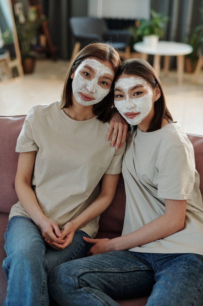 Two young affectionate brunette females with clay mask on faces sitting on couch close to one another in front of camera and looking at you - Photo, Image
