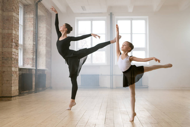 Молодая женщина и маленькая девочка стоят вместе в одной балетной позе и танцуют в танцевальной студии - Фото, изображение