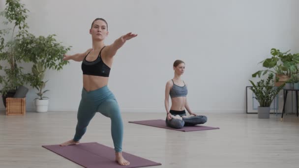 Ευρεία βολή της νεαρής εμφανίσιμης αθλήτριας κάνει ασκήσεις τεντώματος σε λαστιχένιο χαλί, ενώ η γυναίκα ξανθά μαλλιά yogi διαλογίζεται στο παρασκήνιο - Πλάνα, βίντεο