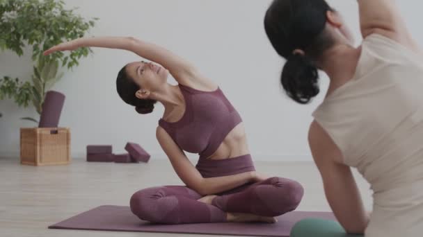 Rear view of attractive sporty Caucasian woman sitting on rubber mat and having personal stretching trainer with female coach sitting in front of her - Footage, Video