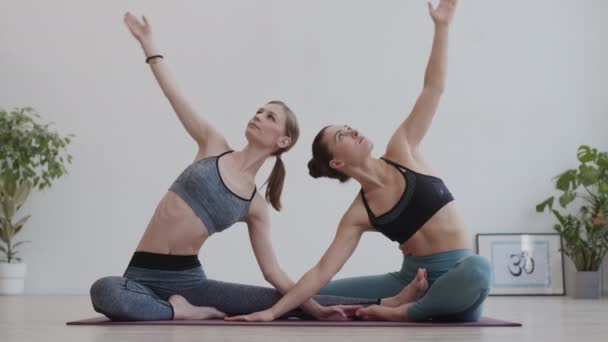 Широкий снимок двух молодых красивых спортсменок в спортивной одежде, занимающихся сложной позой для йоги, сидя на резиновом коврике в студии йоги - Кадры, видео
