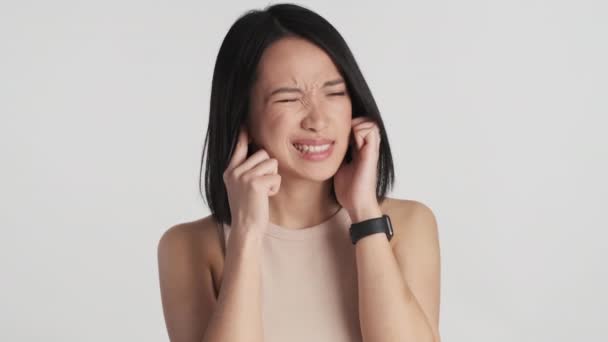 Jonge Aziatische vrouw bedekt oren met vingers omdat het zo luid rond over witte achtergrond. Ik wil het niet horen. - Video