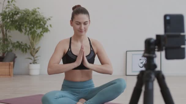 Aussperrung einer jungen kaukasischen Joggerin in Sportbekleidung, die in Lotus-Pose auf einer Yogamatte im Haus vor der Kamera sitzt und ihre Yoga-Praxis auf Video filmt - Filmmaterial, Video