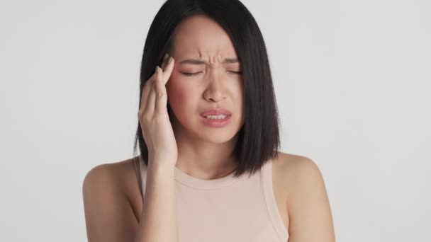 Portrait of upset Asian girl showing headache over white background. Young woman feeling pain - Footage, Video