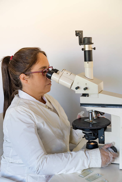 wetenschapper met haar witte schort zittend op een stoel met haar rode bril in haar laboratorium een monster observerend door een microscoop - Foto, afbeelding
