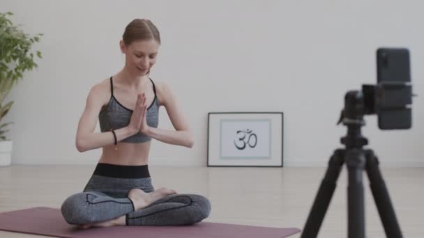 широкая съемка кавказской женщины в спортивной одежде, сидящей в позе лотоса на коврике для йоги в студии йоги перед камерой и снимающей свою практику йоги на видео - Кадры, видео