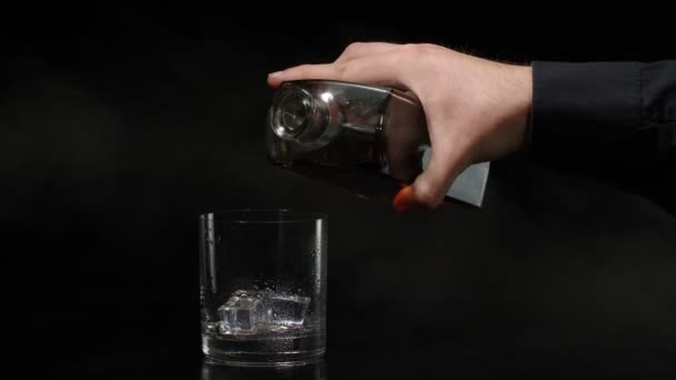 バーマンはウイスキー、コニャック、ブランデーをボトルからグラスに黒の背景にアイスキューブで注ぐ - 映像、動画