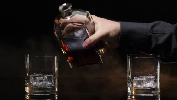 Barman versare acquavite di cognac di whisky dorato dalla bottiglia in bicchieri con cubetti di ghiaccio su sfondo scuro - Filmati, video