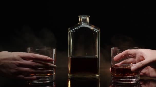 Due mani con bicchieri di whisky al cognac con cubetti di ghiaccio che fanno brindisi su sfondo nero - Filmati, video