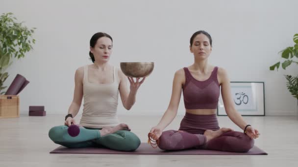 Weitwinkelaufnahme von zwei weiblichen Yogis, die im Innenraum in Lotusposition sitzen und mit geschlossenen Augen meditieren, während sie den Klängen der Yoga-Klangschale lauschen, die eine Frau in Händen hält - Filmmaterial, Video