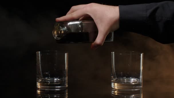 バーマンは金色のウイスキー、コニャック、ブランデー、バーボン、ラム酒を瓶から暗い背景のグラスに注ぐ - 映像、動画